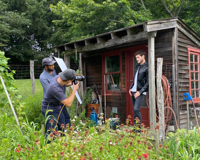 A man poses for a photo outside a rustic wooden shed while two photographers adjust their equipment in a garden filled with flowers.