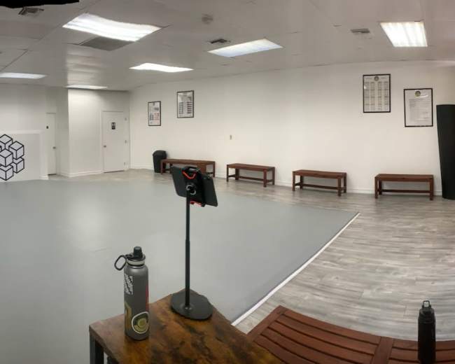 A spacious, empty gym or workout studio with gray flooring, benches along the walls, and exercise equipment in one corner.