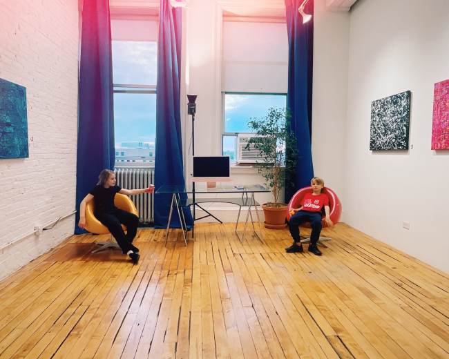 Two people sit in modern chairs facing each other in a bright room with wooden floors and colorful artwork on the walls.