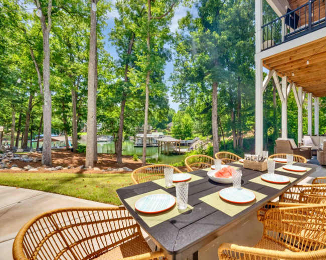 A patio dining area with a large table and woven chairs overlooks a serene waterfront surrounded by trees.