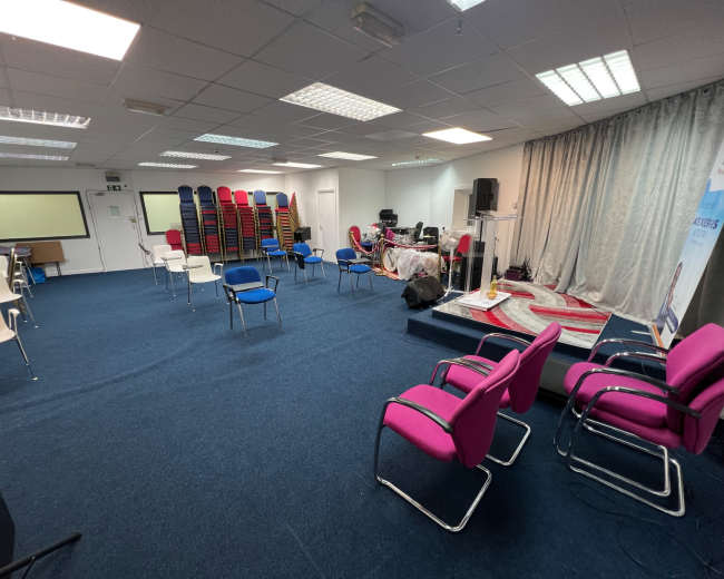 The image shows a conference room arranged with chairs in various colors and stacks of additional chairs against the wall, with a stage setup at the front.