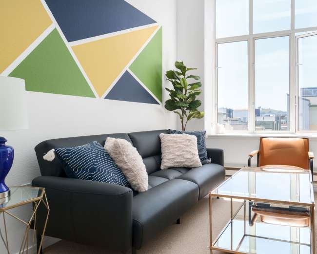 The image shows a modern living room featuring a black sofa with decorative pillows, a geometric wall mural, a potted plant, and a glass coffee table beside an orange accent chair.
