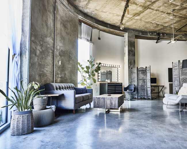 The image shows a spacious industrial-style living area featuring a concrete floor, large windows providing natural light, a gray sofa, a coffee table, indoor plants, and decorative room dividers.