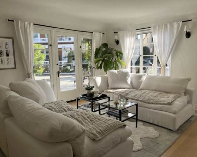 The image shows a bright living room with large windows, white furniture, and a small coffee table adorned with candles and a potted plant.