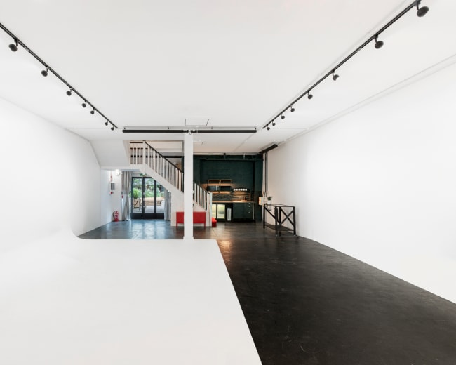 The image shows a spacious, minimalist interior with a staircase on the left, a bar or counter on the right, and large windows at the far end.