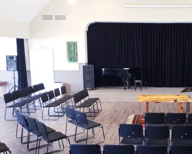 A performance space features rows of black folding chairs facing a wooden table and a grand piano, framed by black curtains and flanked by speakers.