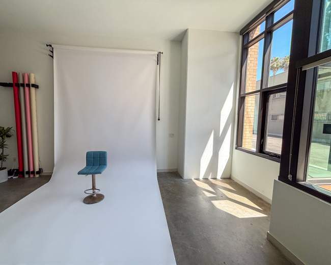 The image shows a minimalist photography studio with a white backdrop, a blue chair on a chrome base, and large windows letting in natural light.