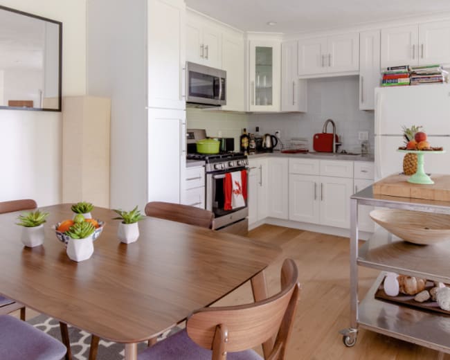 A modern kitchen and dining area feature a wooden table with potted plants, a stainless steel stove, and white cabinetry.