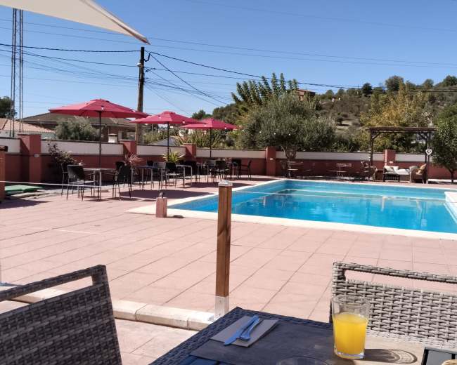 The scene shows a swimming pool surrounded by patio furniture and shaded by umbrellas, with a table set for dining in the foreground.