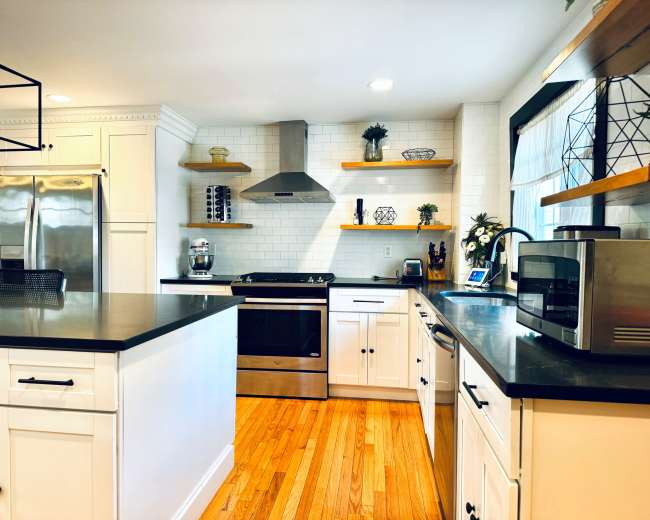 The image shows a modern kitchen with white cabinetry, stainless steel appliances, and wooden shelves displaying decorative items.