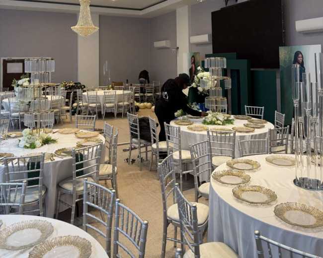 A spacious banquet hall is set up with round tables covered in white tablecloths, elegant chandeliers hanging overhead, and staff preparing the area for an event.