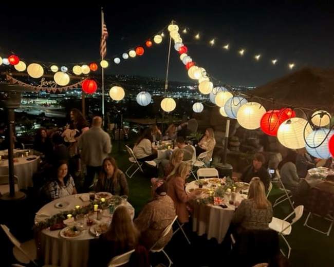 A gathering is taking place outdoors at night, with tables set for guests under colorful lanterns illuminating the scene and a city skyline in the background.