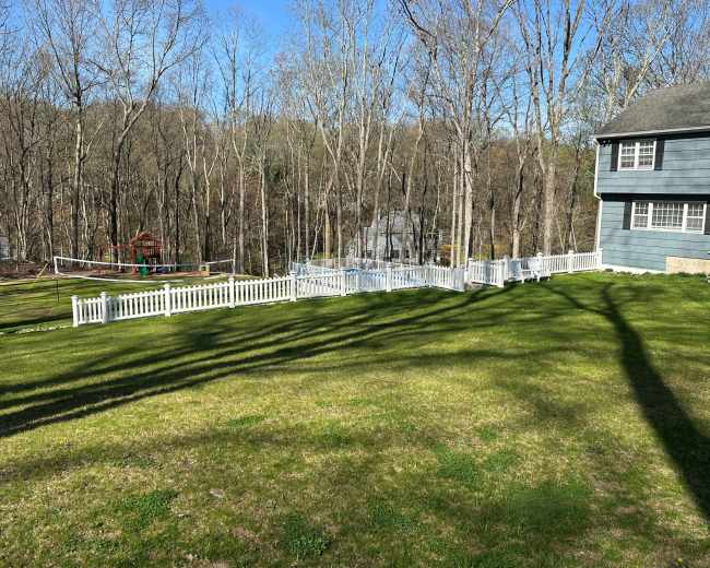 The image shows a backyard with a well-maintained lawn, bordered by a white fence, and trees in the background.