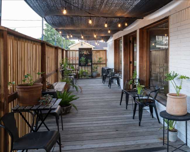 The image shows a wooden deck with potted plants and empty chairs under decorative string lights and a canopy.