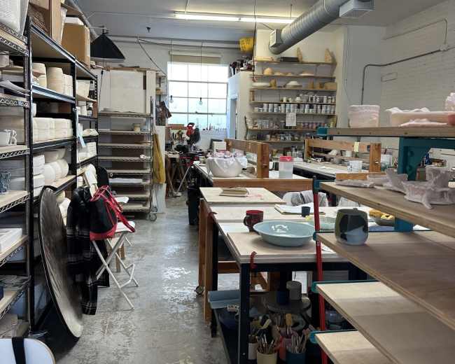The image shows a ceramics studio with shelves filled with pottery supplies, work tables, and equipment arranged in a well-lit space.
