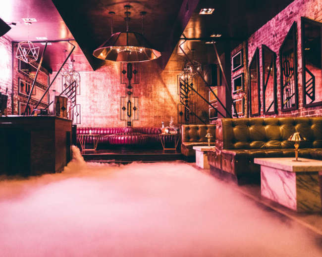 The image shows a dimly lit bar with a foggy floor, showcasing plush seating areas, metallic decor, and geometric light fixtures.