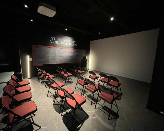 The image shows an empty room set up for a presentation, featuring red folding chairs arranged in rows facing a blank projection screen and a podium.