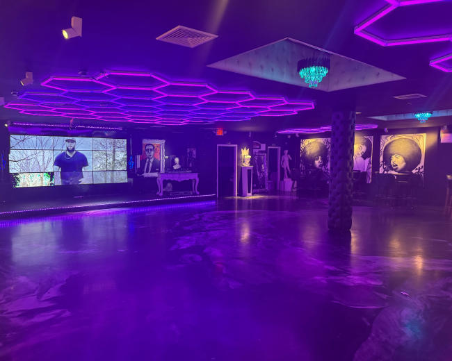 The image shows an illuminated indoor space with purple LED lighting, featuring a large screen and several artworks on the walls.