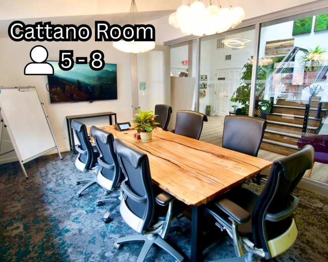 The image shows a conference room with a large wooden table surrounded by black chairs, a whiteboard, and a flat-screen TV mounted on the wall, with a staircase visible in the background.