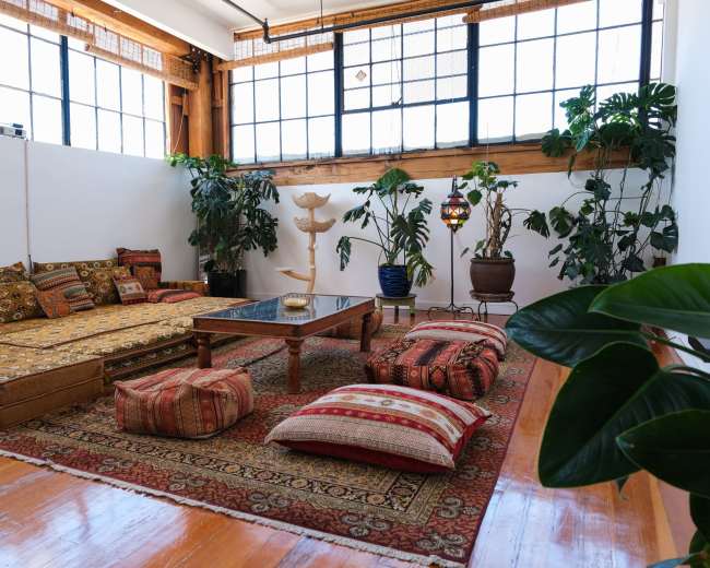 The image features a spacious living area with a low seating arrangement, a central table, and numerous plants, all set against large windows that allow natural light to enter.