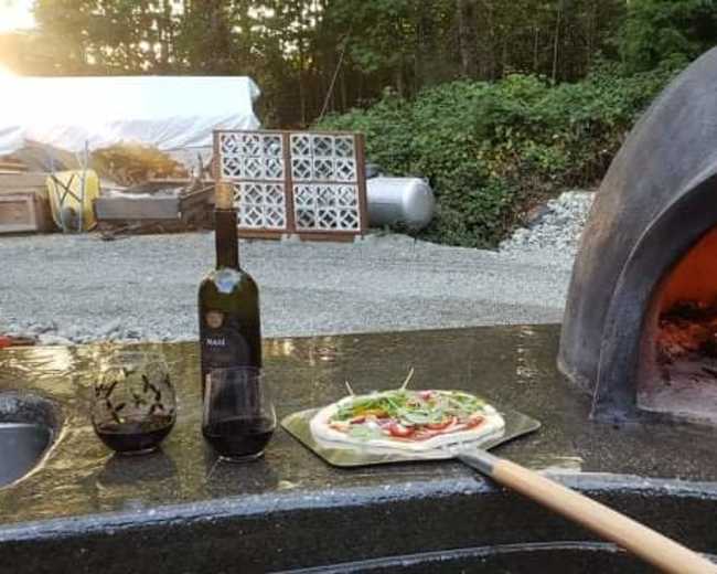 A pizza is being placed into a wood-fired oven with a bottle of olive oil and a glass of wine nearby.