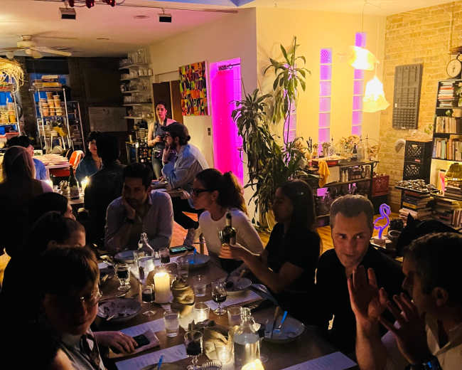 A group of people is seated around a dimly lit dining table, engaged in conversation while candles illuminate the setting.