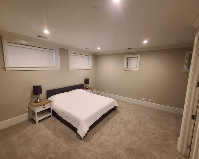 The image shows a spacious bedroom featuring a bed with a white comforter, two bedside tables with lamps, and light-colored walls.