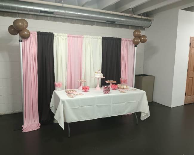 The image shows a decorative table set up with a cake and candy displays, draped with pink, white, and brown curtains in a room with a grey floor and a brown door in the background.