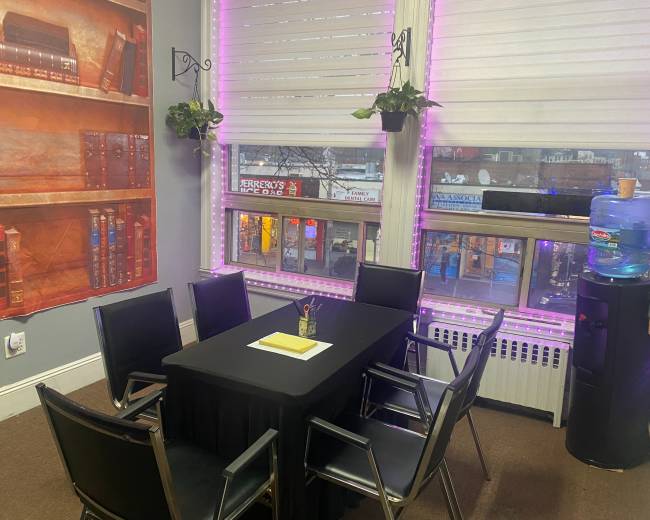 A modern meeting room features a black table surrounded by six chairs, with large windows displaying a street view and shelves decorated with books.
