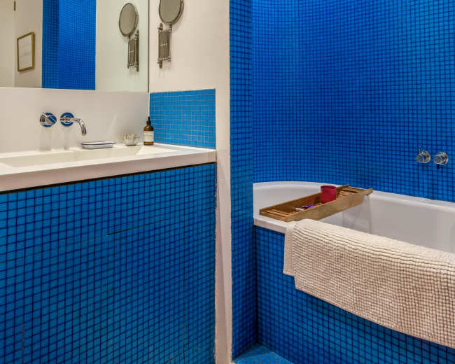 The image shows a modern bathroom with blue mosaic tile walls, a white countertop with sink fixtures, and a bathtub lined with a wooden tray.