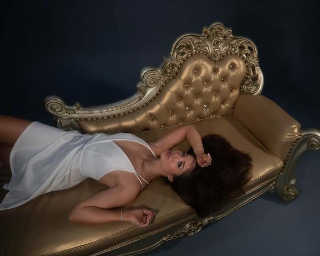 A woman in a white dress lies on a gold tufted chaise lounge against a dark background.