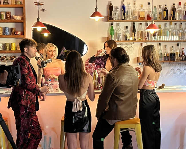 A group of six individuals is standing around a brightly lit bar, engaging in conversation while holding drinks.