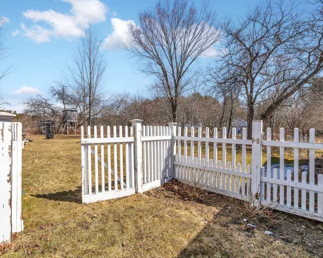 A white picket fence encloses a grassy yard with bare trees and a playset in the background under a clear blue sky.