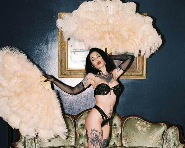 A woman poses with two large feather fans while standing in front of a vintage couch in a dimly lit room.