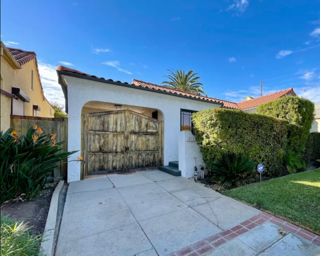 A single-story house with a wooden front door, surrounded by manicured hedges and palm trees, sits on a paved driveway under a blue sky.