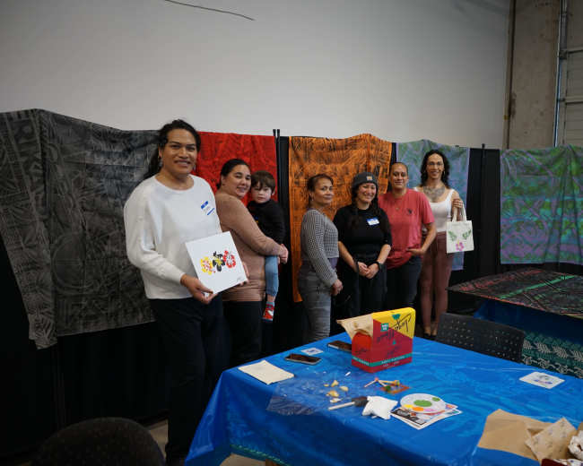 A group of women and children pose together in a room decorated with colorful fabric displays and art supplies on a table.