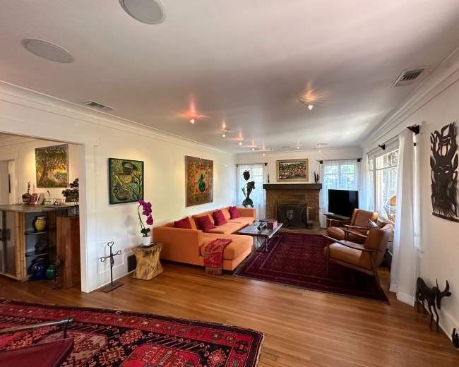 The image shows a spacious living room featuring an orange sectional sofa, wooden furniture, colorful artwork on the walls, and a fireplace.