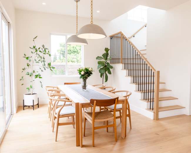 A spacious dining area features a wooden table surrounded by chairs, a vase of flowers in the center, and a staircase leading to an upper level.