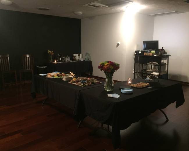 A room set up for a gathering, featuring two tables with food and drinks, surrounded by dark walls and hardwood flooring.