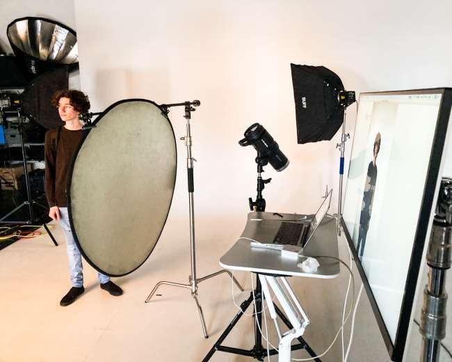 A male model stands beside a large reflector in a photography studio equipped with lighting and a computer displaying a live image.