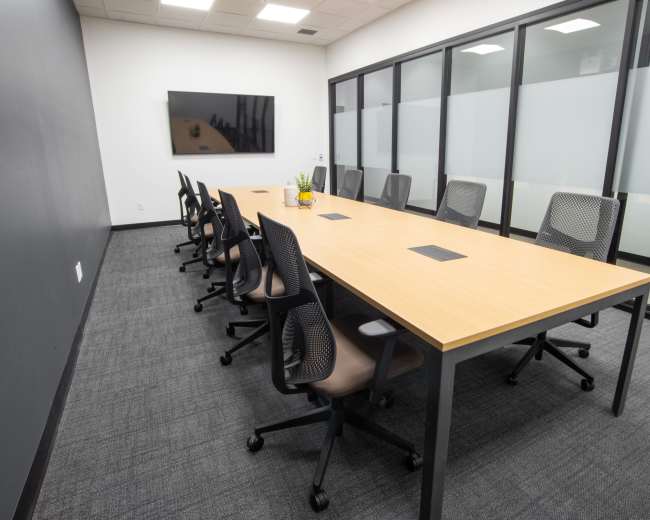 The image shows a modern conference room with a long wooden table surrounded by ergonomic chairs and a wall-mounted screen.