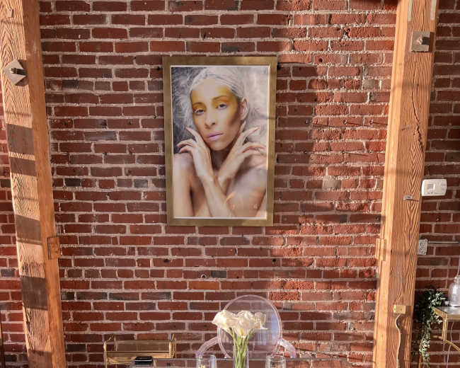 A small silver table is placed on a gray rug in front of a brick wall, adorned with a framed portrait of a person hanging above it.