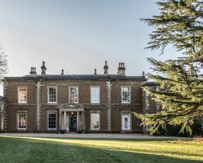 The image shows a large, two-story brick house with a symmetrical facade, featuring a central entrance and surrounded by a manicured lawn and trees.