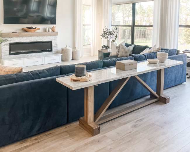 A modern living room features a blue sofa, a marble-top console table, a mounted television, and large windows covered with light curtains.