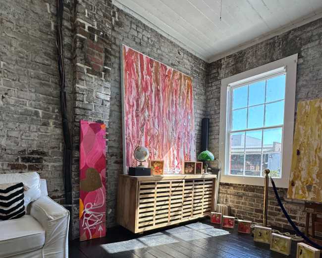 The image shows a room with exposed brick walls, featuring colorful paintings, a wooden console table, and a white sofa.