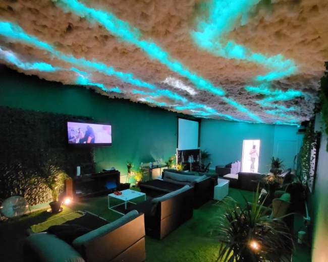 The image shows a modern lounge area with plush sofas, a television on the wall, and ambient lighting created by green and blue hues, alongside various plants and a textured ceiling resembling clouds.
