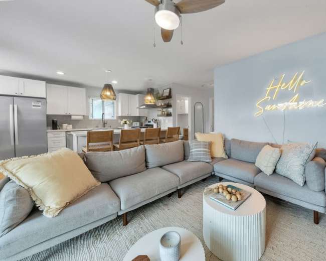 The image shows a modern living room featuring a gray sectional sofa, a coffee table, decorative cushions, and a wall-mounted neon sign with the phrase "Hello Sunshine."