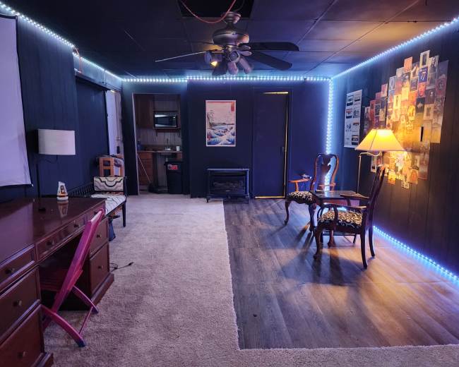 A room with a carpeted floor, featuring a desk with a pink chair, two armchairs, a lamp, a kitchenette area, and walls illuminated by LED strip lighting.