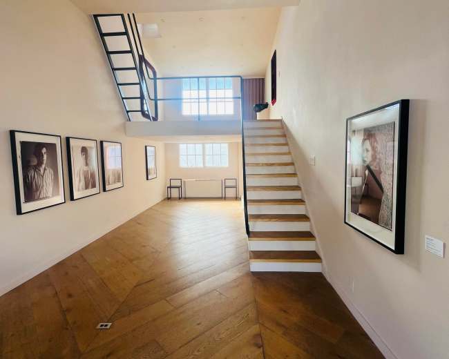The image shows a spacious gallery with wooden flooring, featuring portraits on the walls and a staircase leading to the upper level.