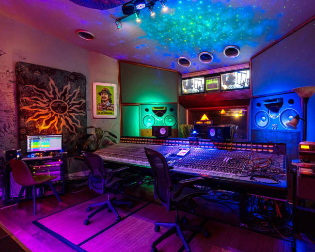 A music recording studio with a large mixing console, colorful lighting, and various audio equipment arranged around the space.